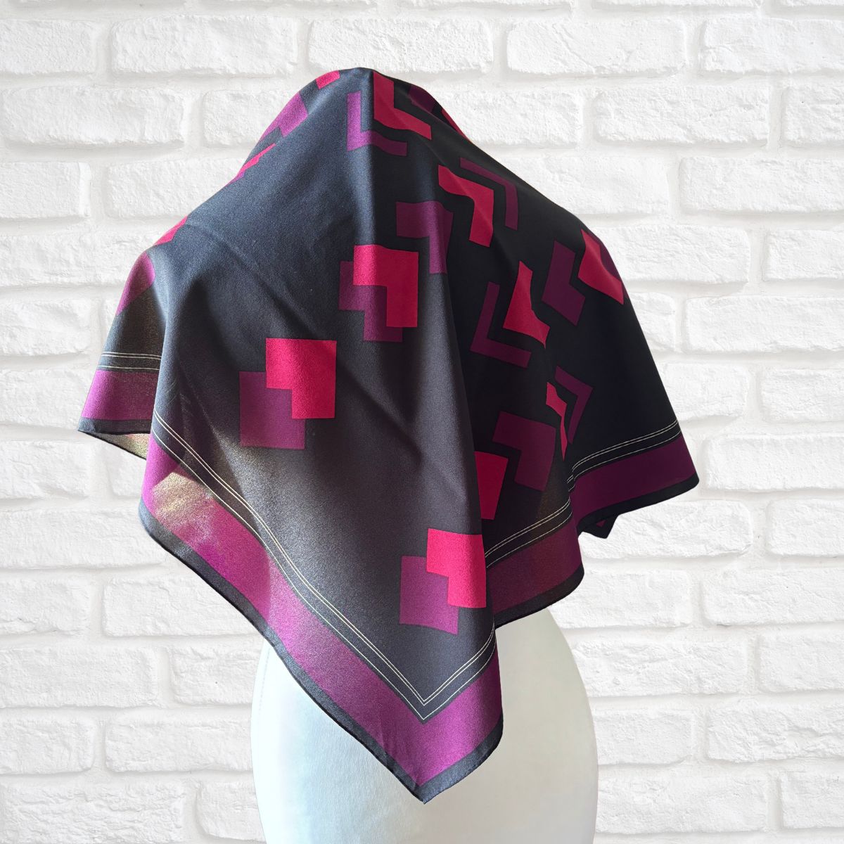 Black scarf with pink geometric patterns draped over a mannequin against a white brick wall.