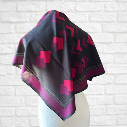 Black scarf with pink geometric patterns draped over a mannequin against a white brick wall.
