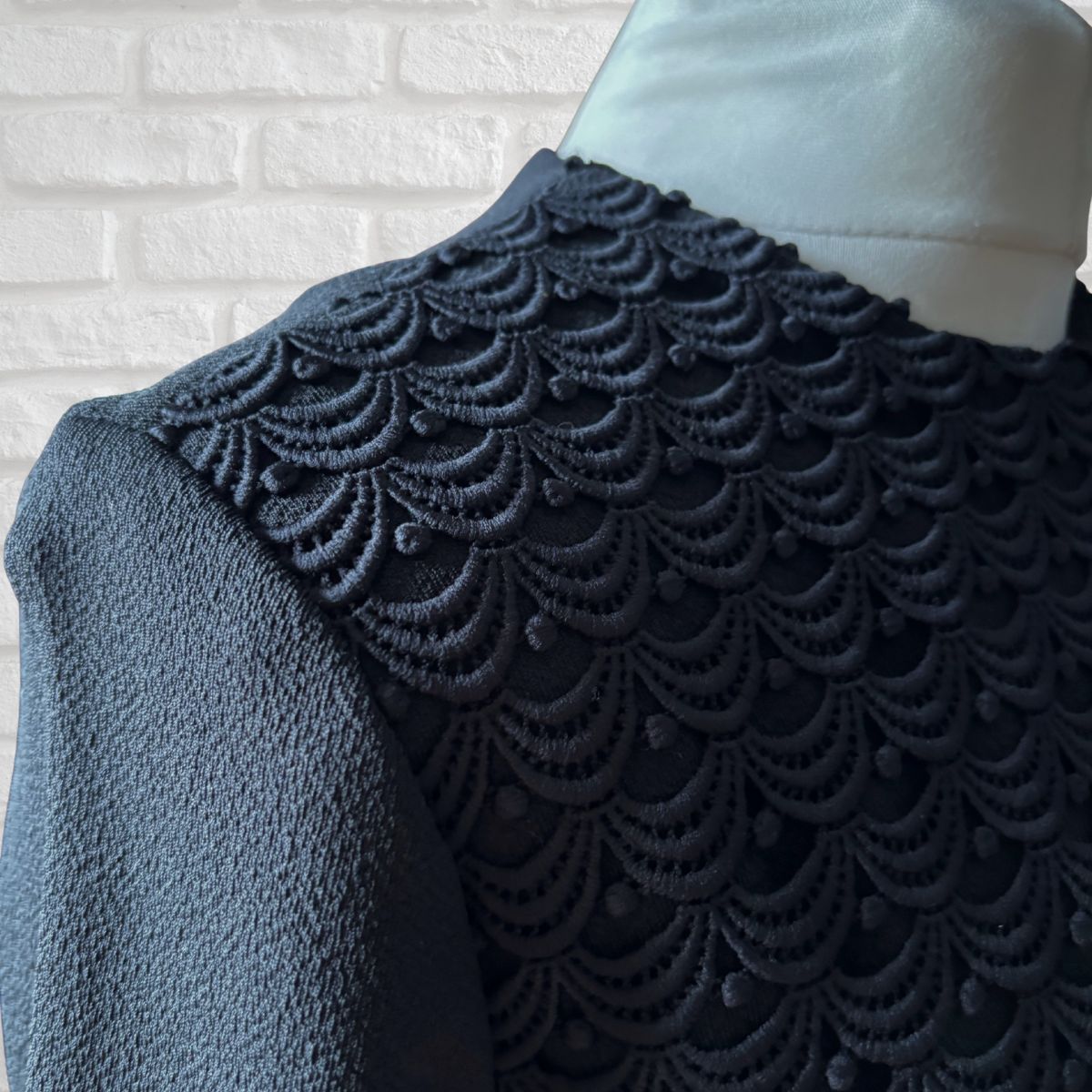 Textured fabric with scalloped edge on a mannequin against a white brick wall.