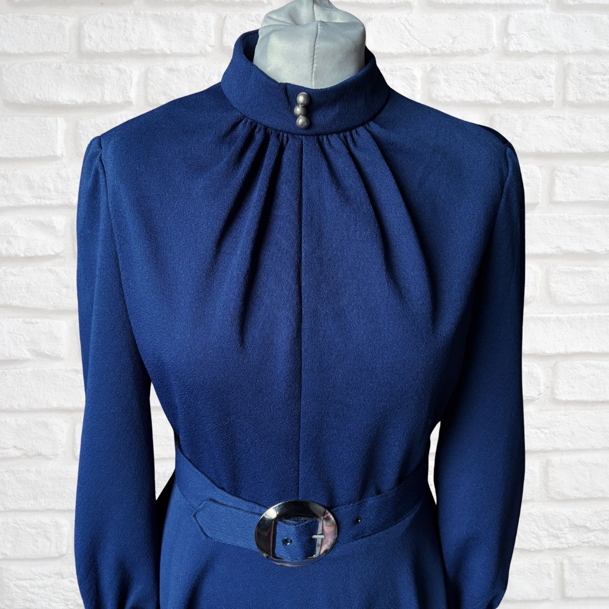 Blue dress with a high collar and belt on a mannequin against a white brick wall.
