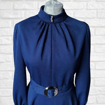 Blue dress with a high collar and belt on a mannequin against a white brick wall.