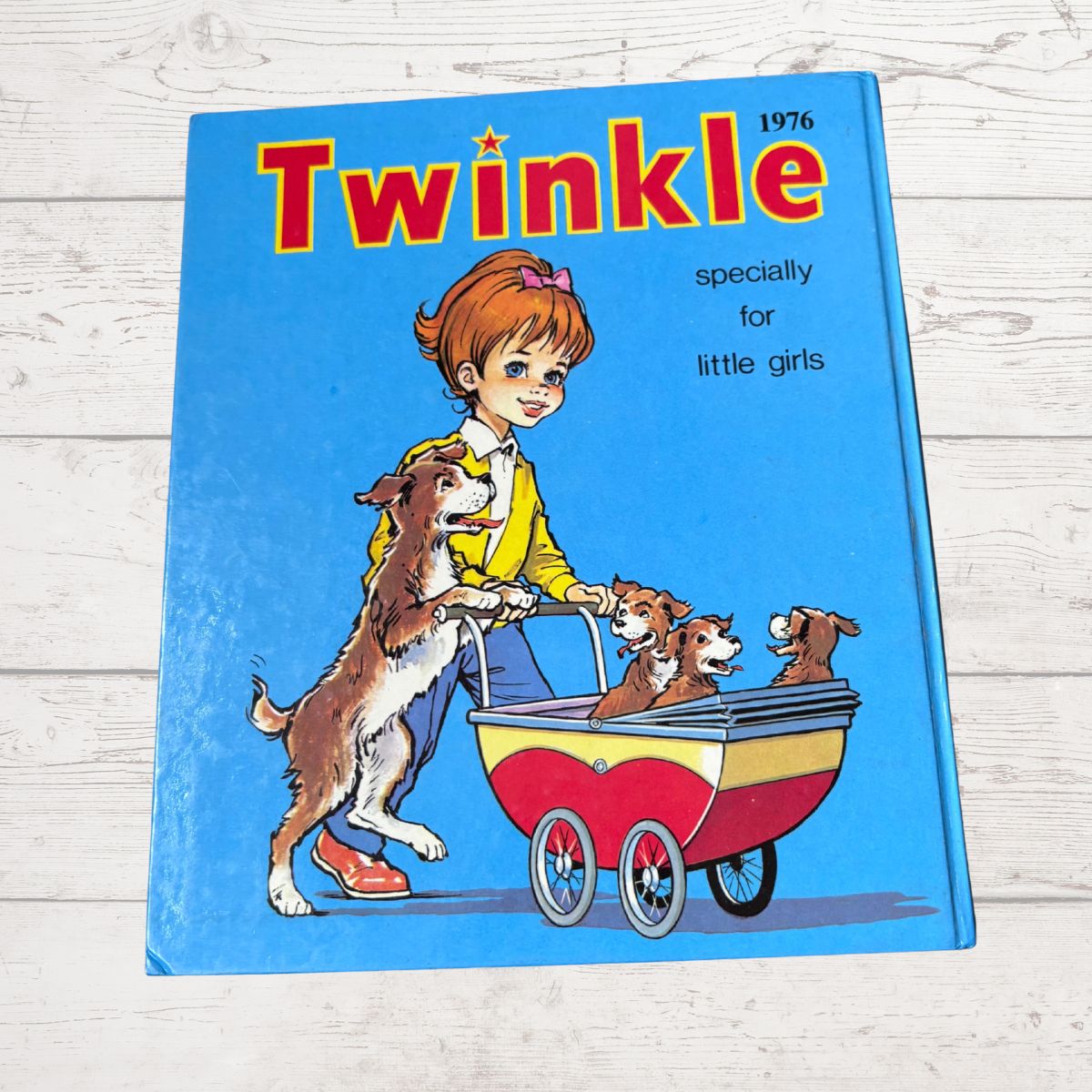 A vintage children's book titled 'Twinkle 1976' featuring an illustration of a girl in a yellow jacket pushing a red and cream baby stroller with three dogs, set against a light wood background.