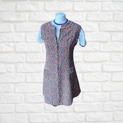 A short-sleeve floral-patterned apron dress displayed on a light blue mannequin torso against a white brick wall background.
