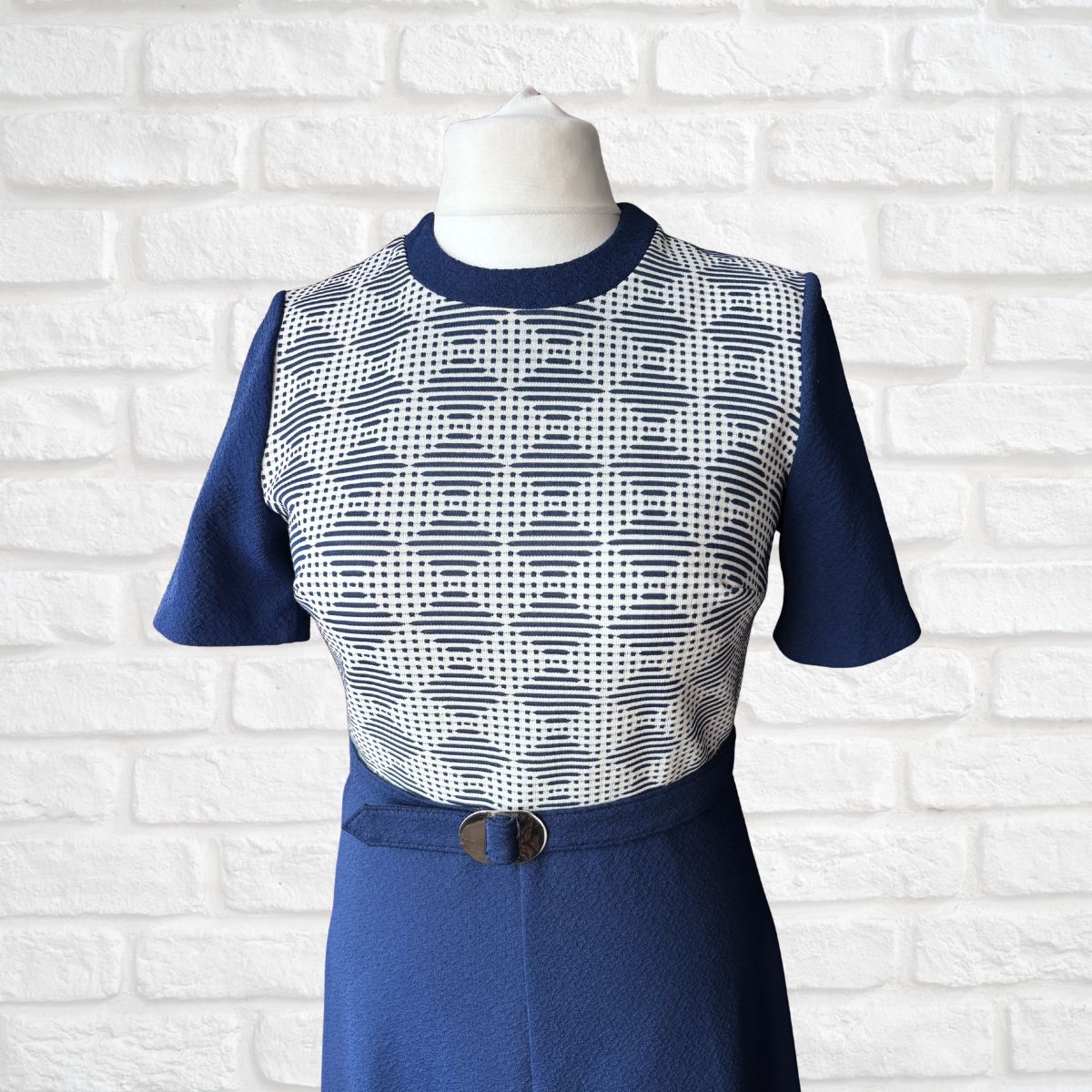 Mannequin wearing a blue and white patterned dress against a white brick wall.