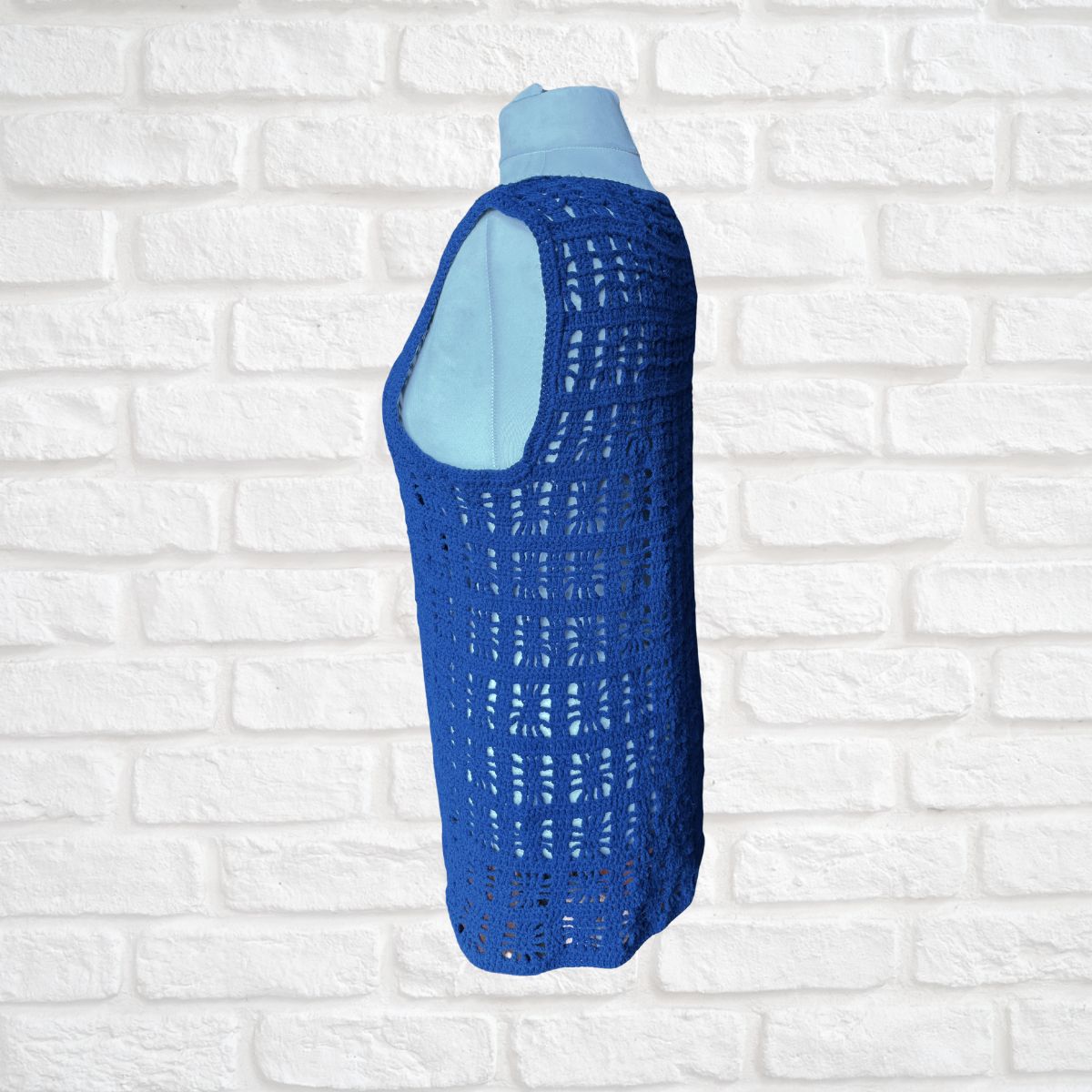 A sleeveless blue crochet vest displayed on a light blue mannequin torso against a white brick wall background.