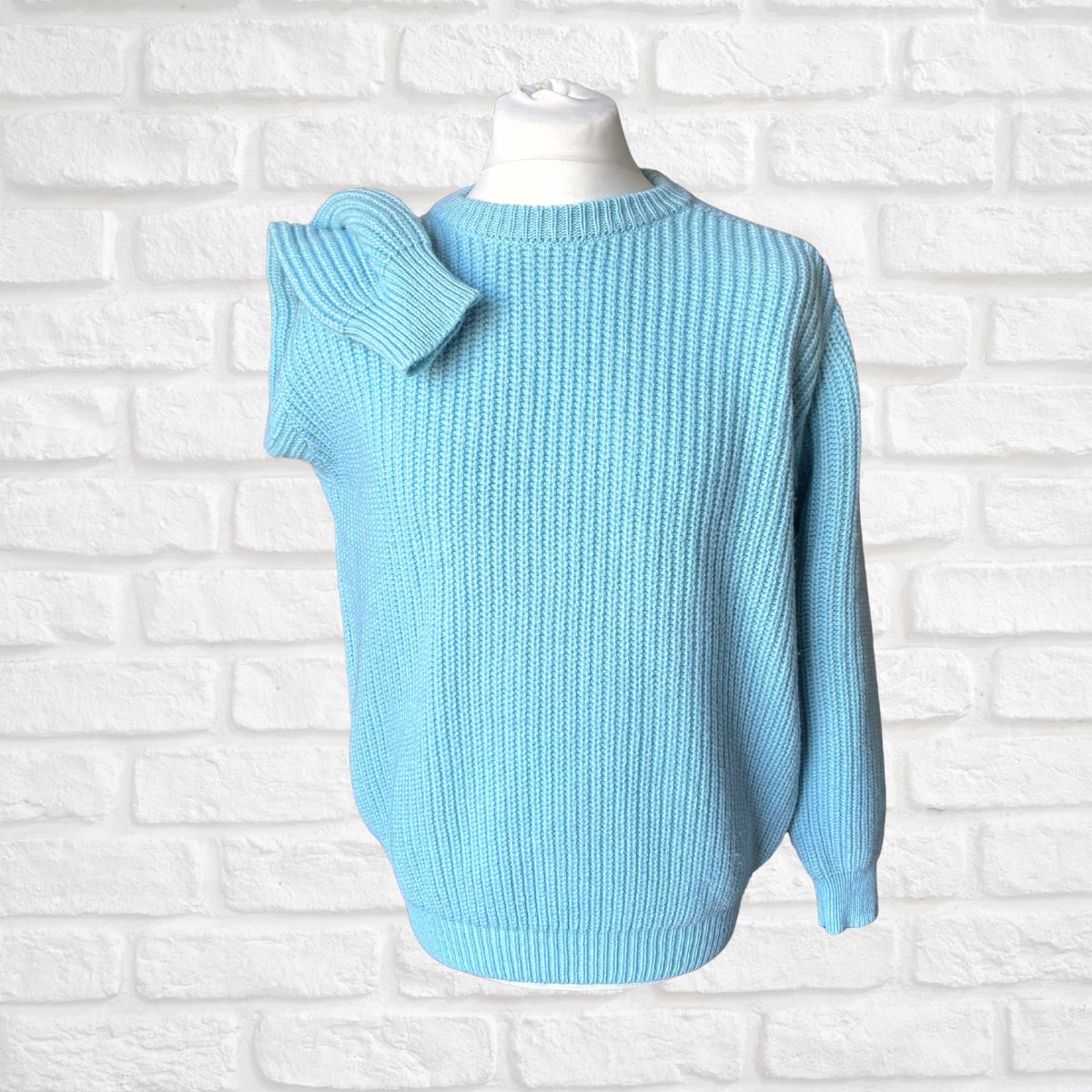 A light blue ribbed knit sweater displayed on a white mannequin torso against a white painted brick wall background.