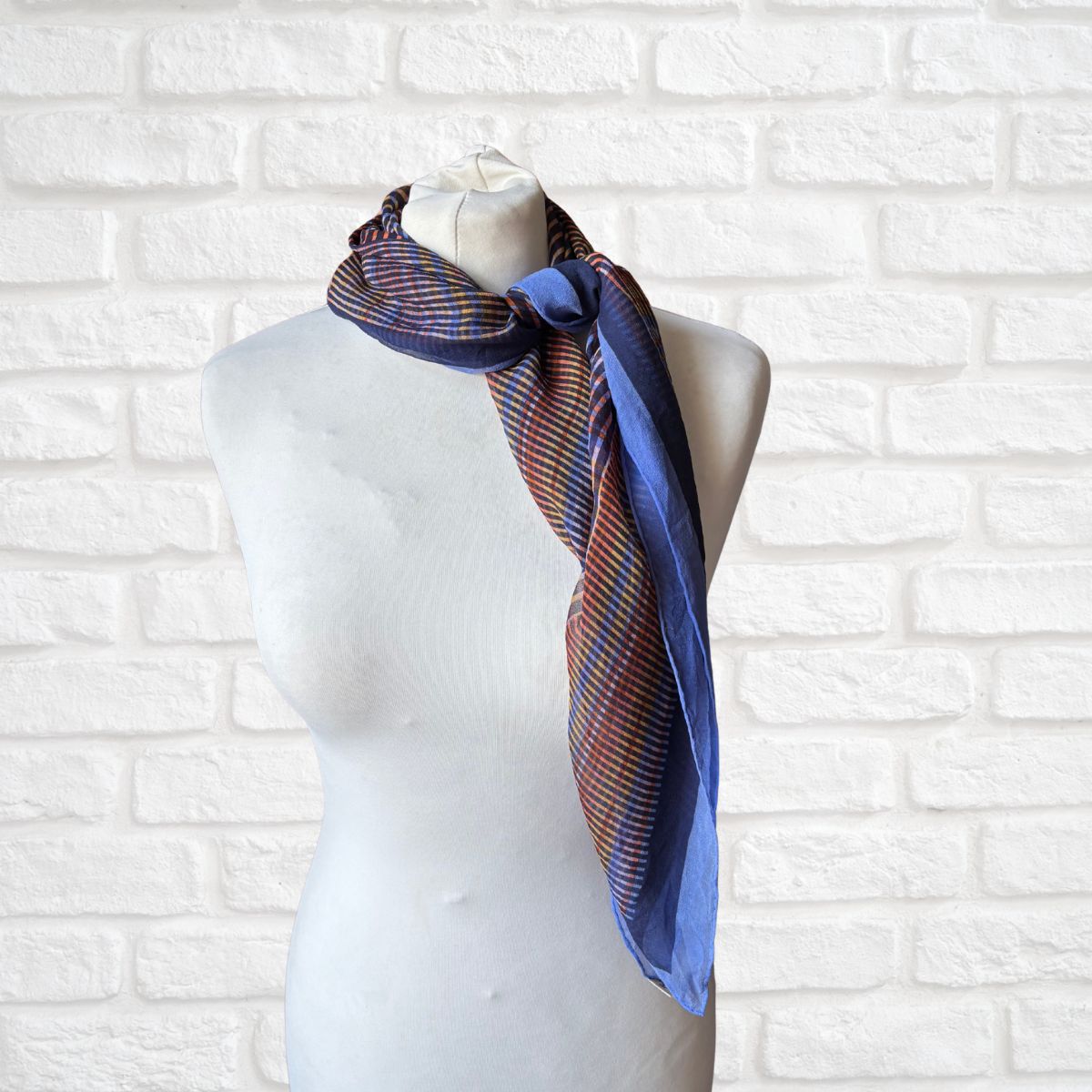 Mannequin wearing a blue and orange scarf against a white brick wall.