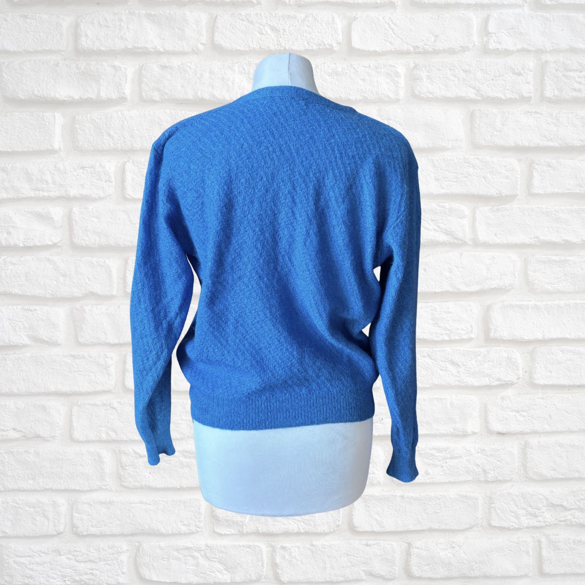 A bright blue knit sweater displayed on a white mannequin torso, shown from the back against a white brick wall background.