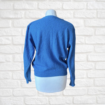 A bright blue knit sweater displayed on a white mannequin torso, shown from the back against a white brick wall background.