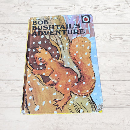 Vintage children's book titled 'Bob Bushtail's Adventure' featuring an illustration of a squirrel on a tree trunk, photographed on a light wood plank surface.
