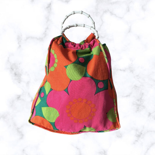 A fabric bag  with a bold mid-century modern floral pattern in pink, orange, green, and purple, featuring a silver-toned bamboo-style top handle, set against a white marble background.