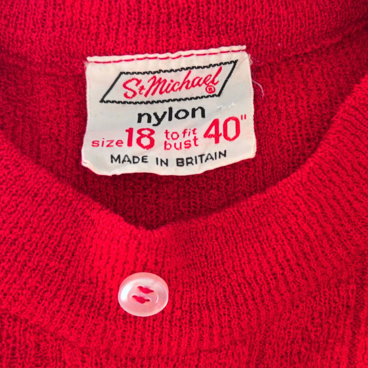 Close-up of a red knit cardigan sweater showing the inner neck label which reads 'St. Michael nylon size 18 to fit bust 40" MADE IN BRITAIN' and a single light pink button at the neckline.