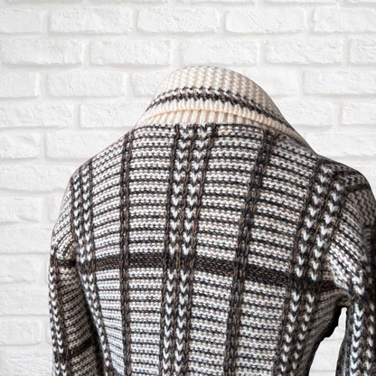 A close-up of the back of a knit cardigan sweater featuring a cream, brown, and black textured pattern, displayed against a white brick wall.
