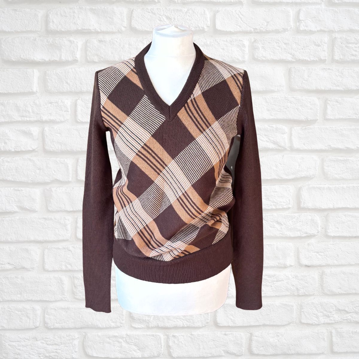 A long-sleeve V-neck sweater displayed on a white mannequin torso against a white brick wall. The sweater features solid dark brown sleeves, collar, and hem, with a plaid yoke panel in brown, beige, and white.