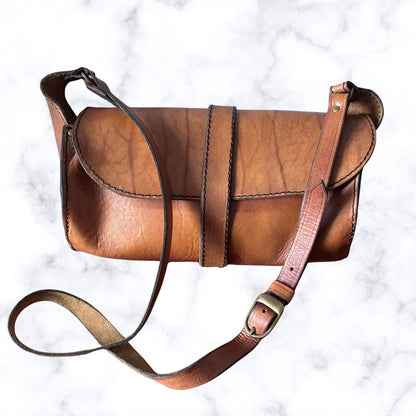 A brown leather shoulder bag with a long strap and brass buckle, laid flat on a white marble background.