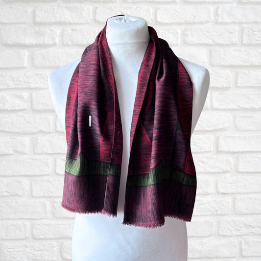 A dark red and black patterned scarf with olive green bands and fringed edges, draped on a white mannequin torso against a white brick wall background.