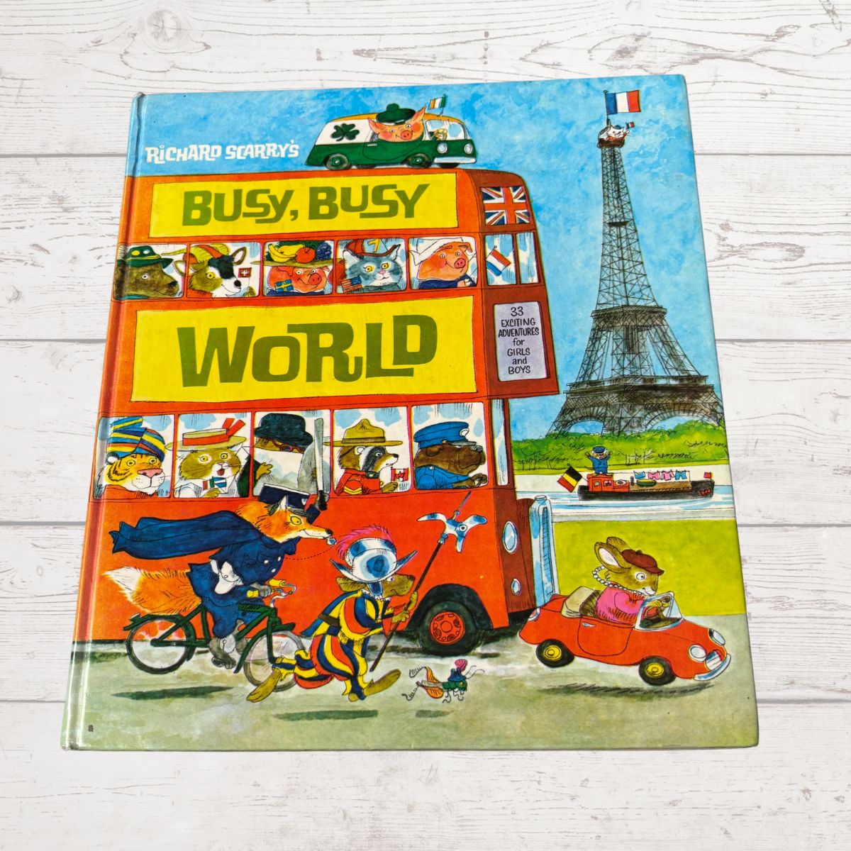 Book cover of 'Busy, Busy World' with a colorful illustration of a bus and Eiffel Tower on a wooden surface.