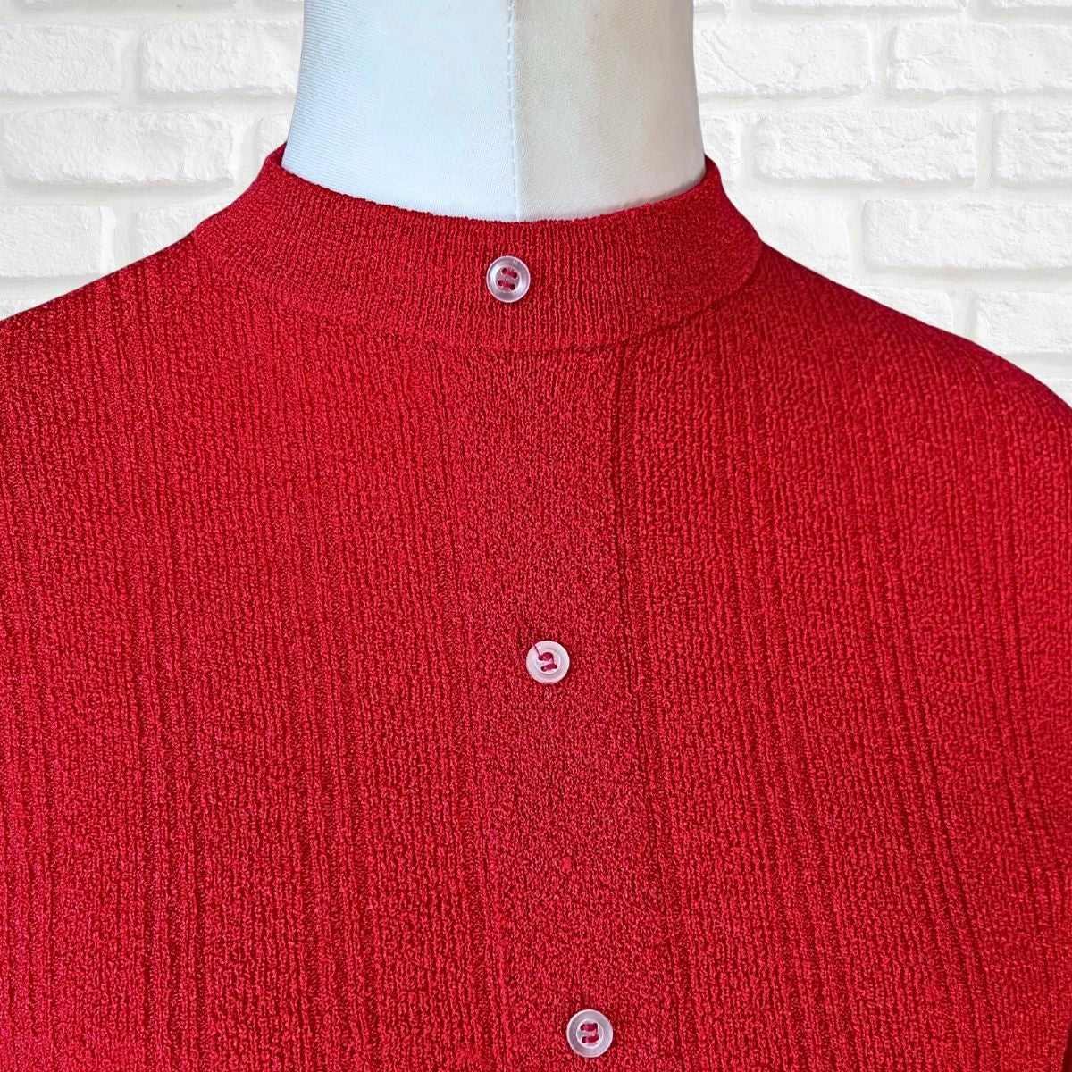 A close-up of a red ribbed knit cardigan sweater displayed on a white mannequin torso, featuring a mock neck and three light pink buttons, set against a white brick wall background.