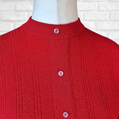 A close-up of a red ribbed knit cardigan sweater displayed on a white mannequin torso, featuring a mock neck and three light pink buttons, set against a white brick wall background.