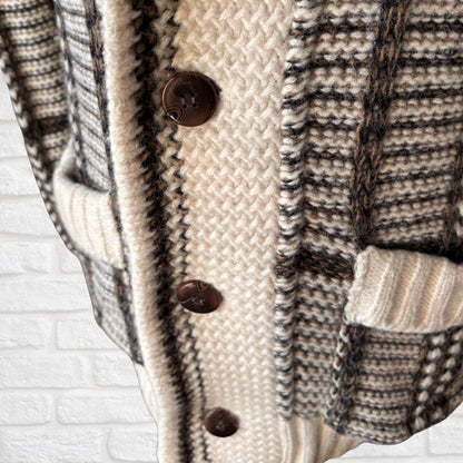 Close-up of a knit cardigan sweater with a cream and black-and-beige plaid pattern, featuring dark brown buttons and ribbed trim, set against a white brick wall background.