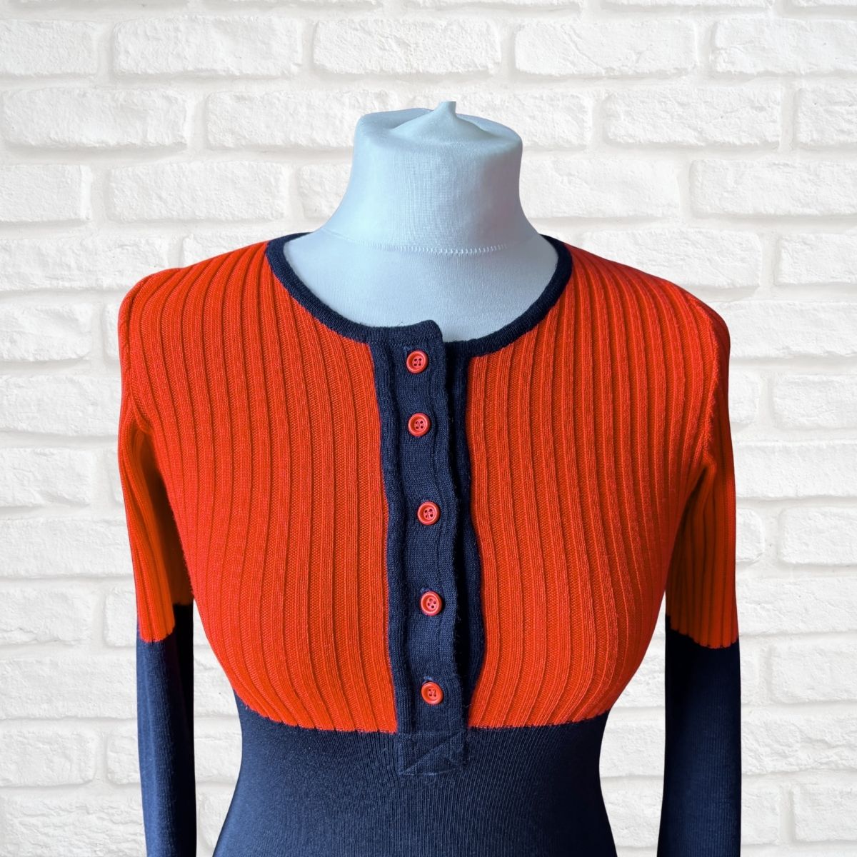 A colour block ribbed knit sweater displayed on a mannequin torso against a white brick wall background. The cardigan features a vibrant orange-red body, contrasting dark navy blue collar and button placket, and navy blue sleeves.