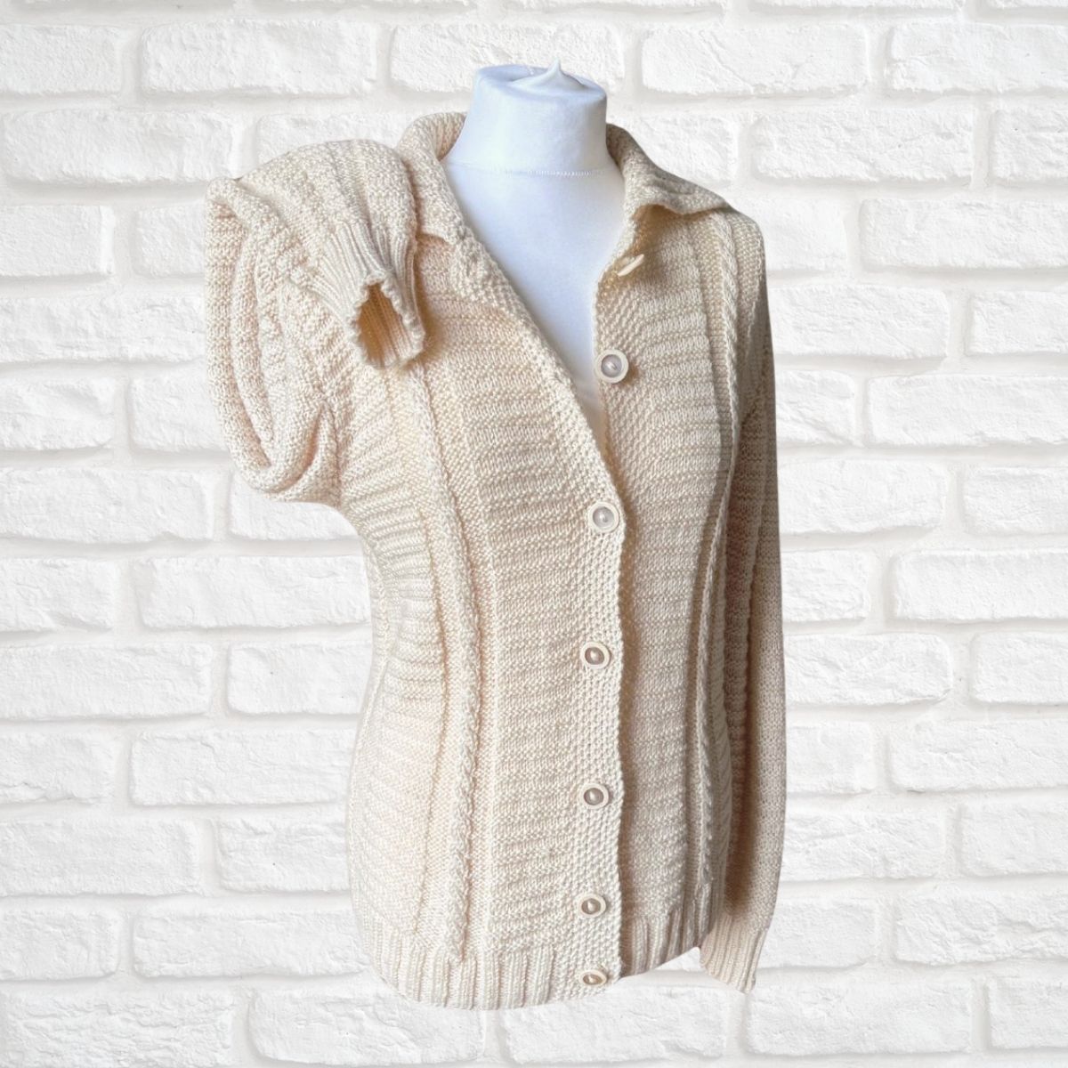 A cream-colored chunky knit cardigan sweater with a short puff sleeve, button-front closure, and ribbed collar, displayed on a white mannequin torso against a white painted brick wall.