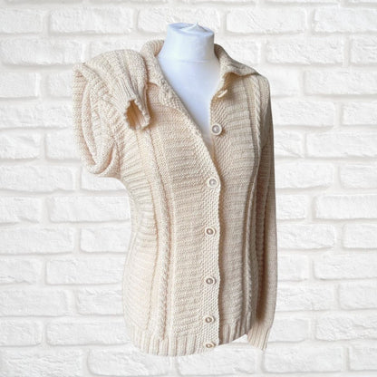 A cream-colored chunky knit cardigan sweater with a short puff sleeve, button-front closure, and ribbed collar, displayed on a white mannequin torso against a white painted brick wall.