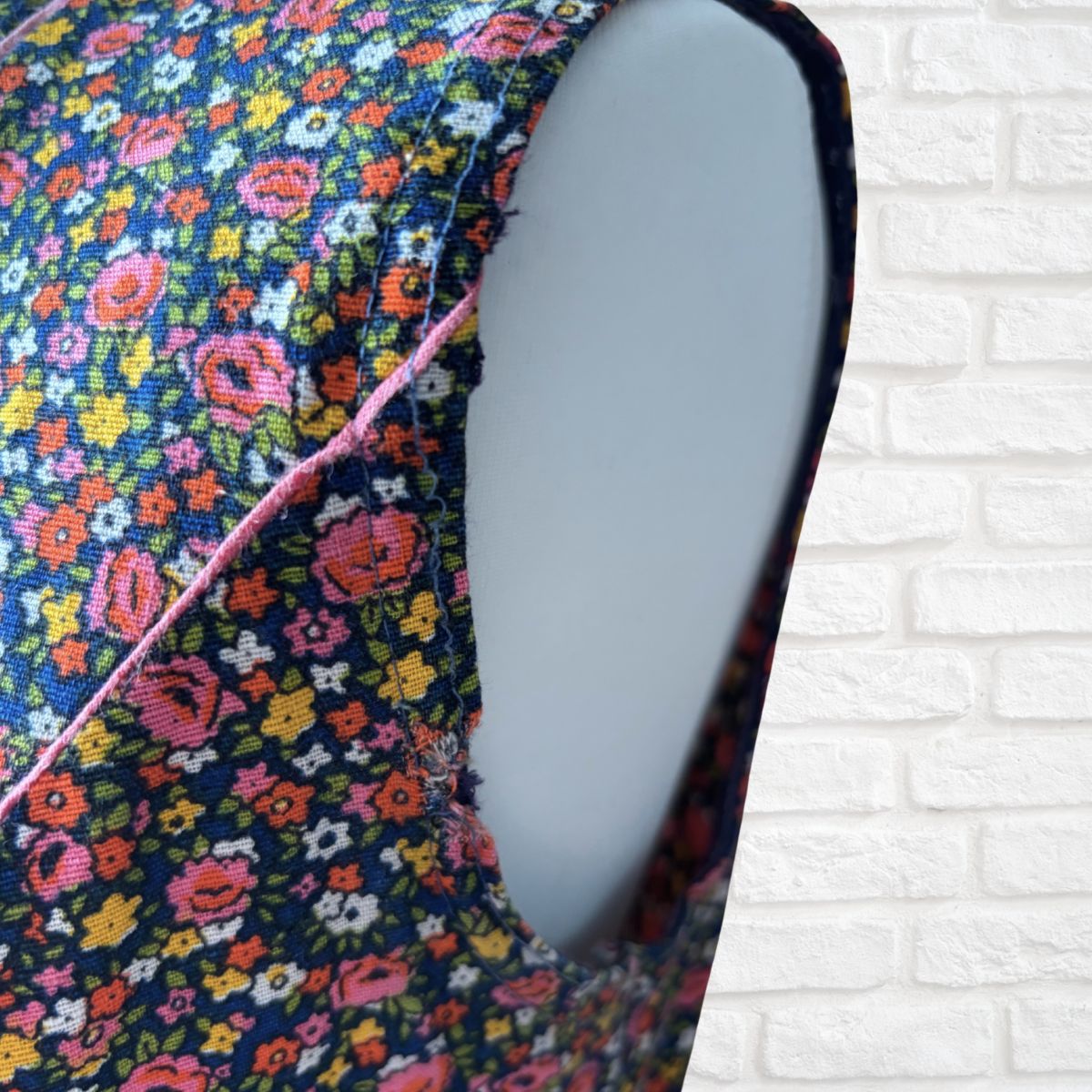A close-up of a sleeveless dress with a dark navy blue background and a dense multicolor floral print, featuring a pink contrast stitch along the side seam. The garment is displayed on a light gray mannequin torso against a white brick wall background.
