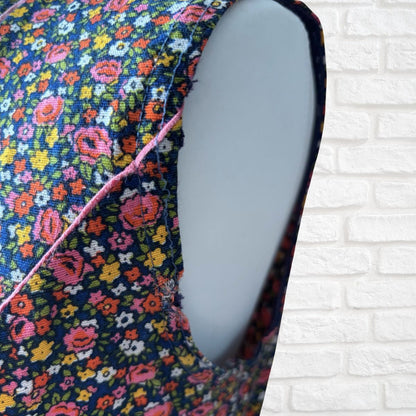 A close-up of a sleeveless dress with a dark navy blue background and a dense multicolor floral print, featuring a pink contrast stitch along the side seam. The garment is displayed on a light gray mannequin torso against a white brick wall background.