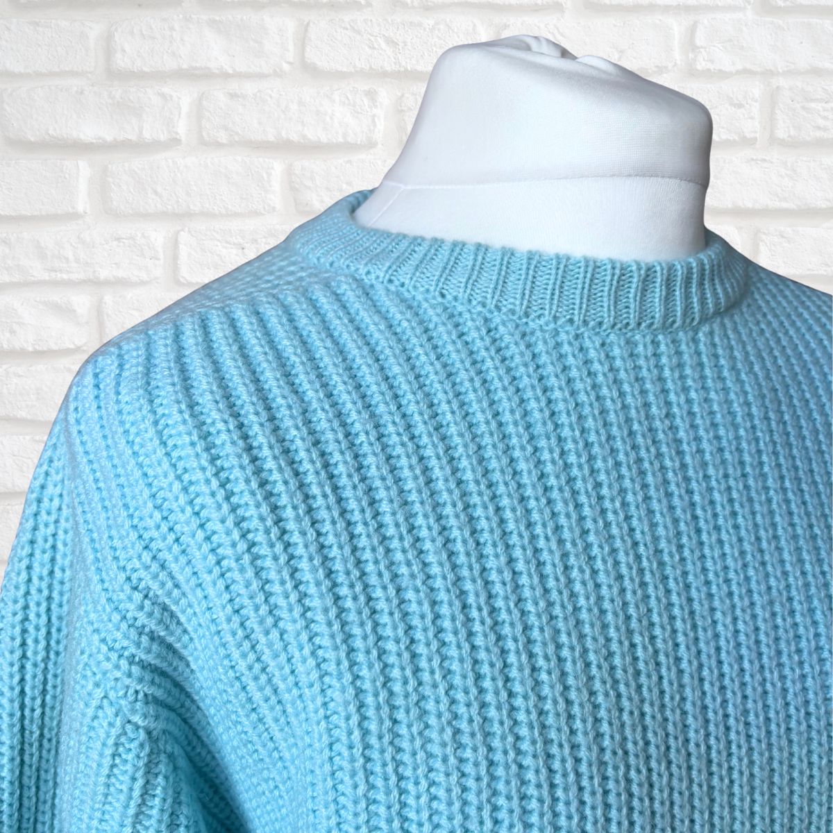 A light blue ribbed knit sweater displayed on a white mannequin torso against a white brick wall background.