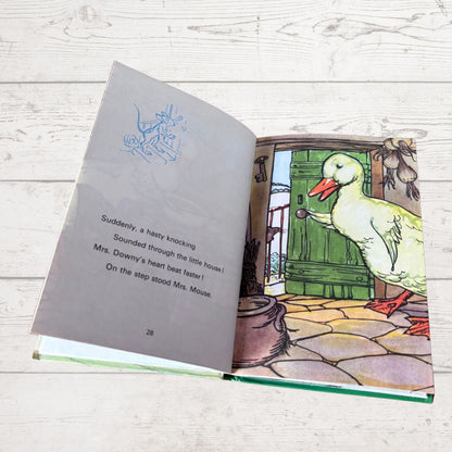 An open children's picture book lying on a whitewashed wooden surface, showing illustrated pages with text and a cartoon duck character standing by a green door.