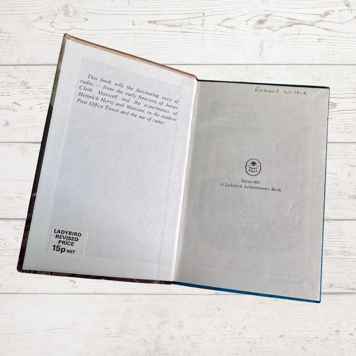 An open book lying flat on a white wooden surface, showing the interior pages with printed text, a small circular logo, and handwritten notes. The book has a dark brown cover edge and a blue spine edge visible.