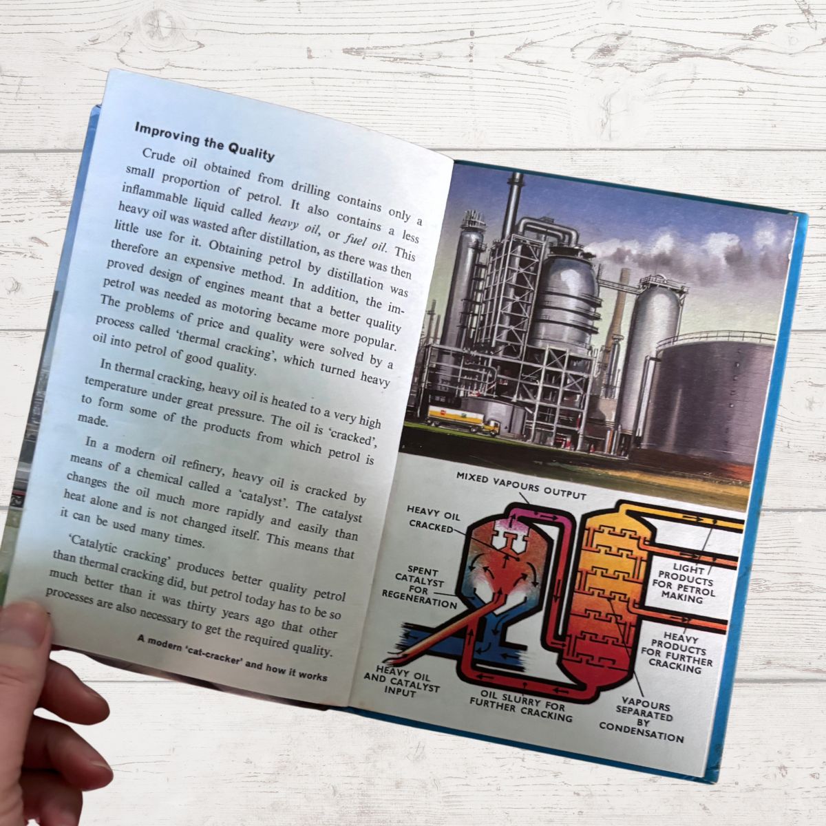 An open printed book page showing text about crude oil refining, with a large industrial refinery photo and a colorful schematic diagram of a refinery process. A hand is holding the page at the lower left, and the background is a light wood surface.