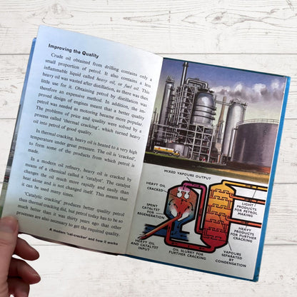 An open printed book page showing text about crude oil refining, with a large industrial refinery photo and a colorful schematic diagram of a refinery process. A hand is holding the page at the lower left, and the background is a light wood surface.