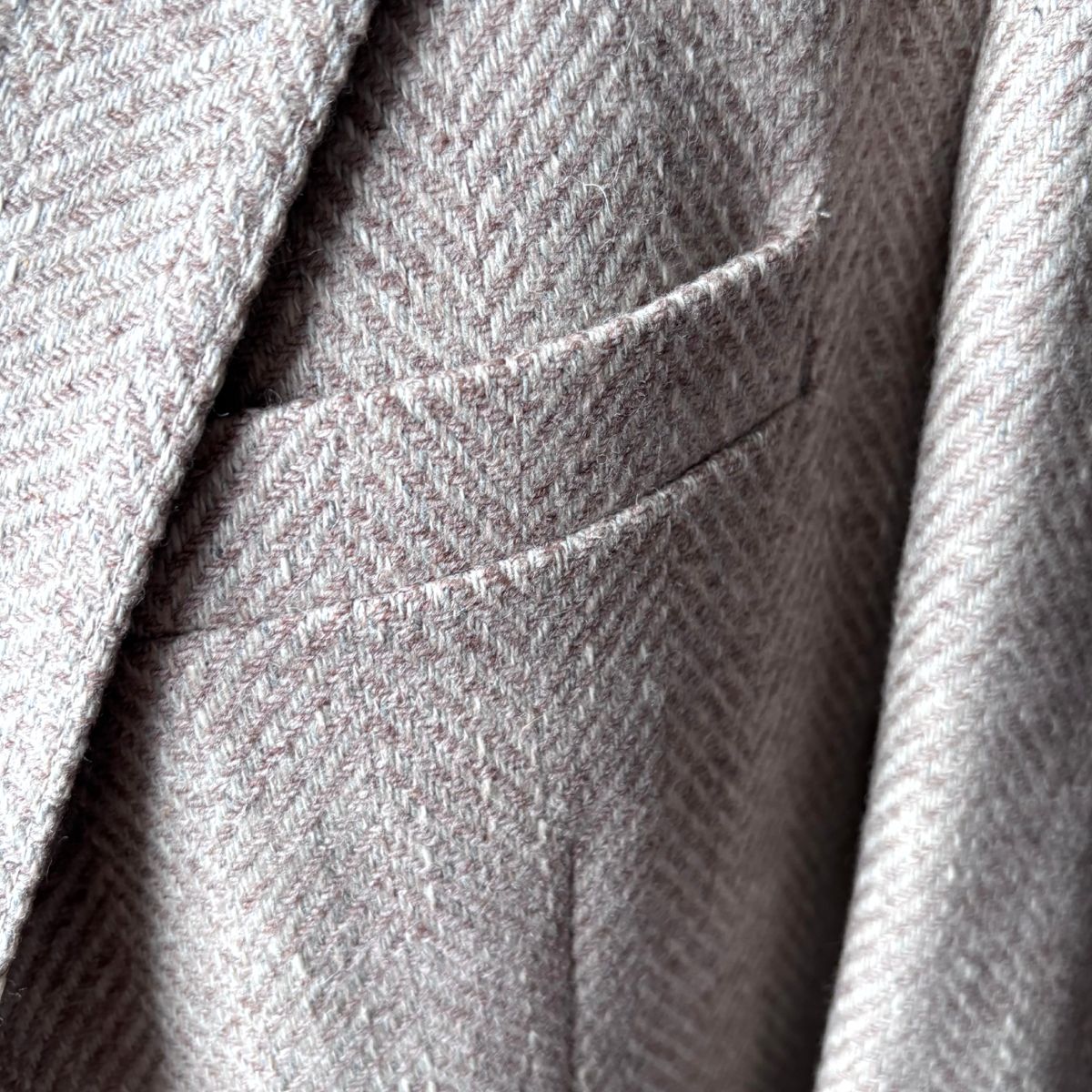 Close-up of a herringbone tweed fabric jacket, showing the texture and a pocket detail in a light gray and beige colorway.
