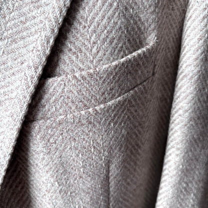 Close-up of a herringbone tweed fabric jacket, showing the texture and a pocket detail in a light gray and beige colorway.