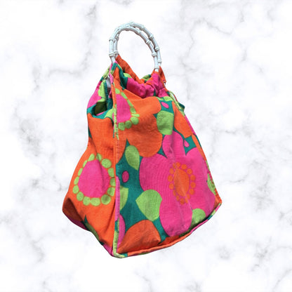 A top handle bag with a vibrant mid-century floral pattern in orange, pink, green, and teal, featuring a white twisted handle, set against a white marble background.