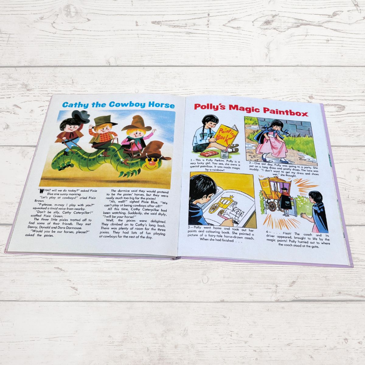 An open children's book spread laid flat on a white wood surface, showing two illustrated pages with colorful comic panels and text. The left page is titled 'Cathy the Cowboy Horse' and the right page is titled 'Polly's Magic Paintbox.