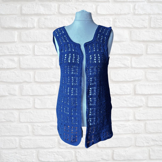 A sleeveless open-knit crochet waistcoat  in a rich royal blue colour, displayed on a light blue mannequin torso against a white painted brick wall background.