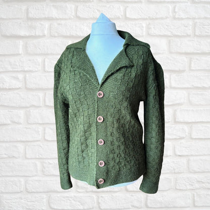A green cable-knit cardigan sweater with a collar and pink buttons, displayed on a light blue mannequin torso against a white brick wall background.