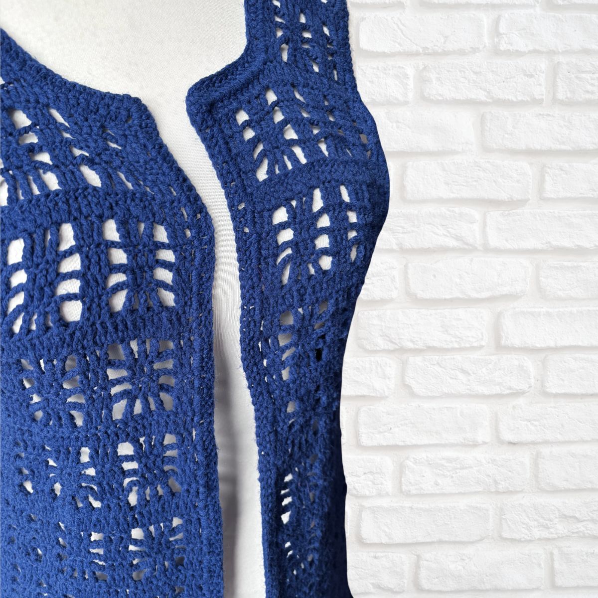 A close-up of a blue crocheted sleeve or trim with an openwork lace pattern, shown against a white brick wall background.