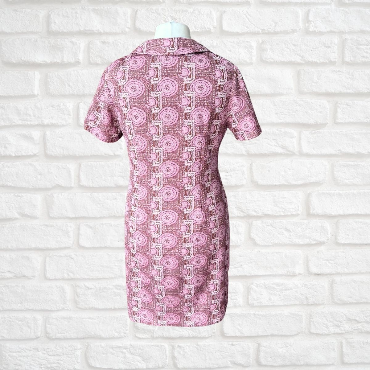 A short-sleeve collared dress displayed on a mannequin torso against a white brick wall. The dress features a pink and white geometric pattern with circular motifs.