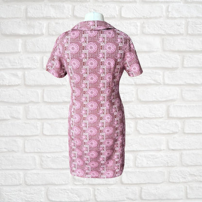 A short-sleeve collared dress displayed on a mannequin torso against a white brick wall. The dress features a pink and white geometric pattern with circular motifs.