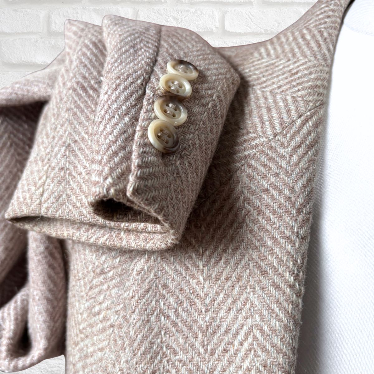 Close-up of a folded tweed jacket sleeve with a herringbone pattern in warm taupe and beige, featuring four cream buttons, set against a white brick wall background.