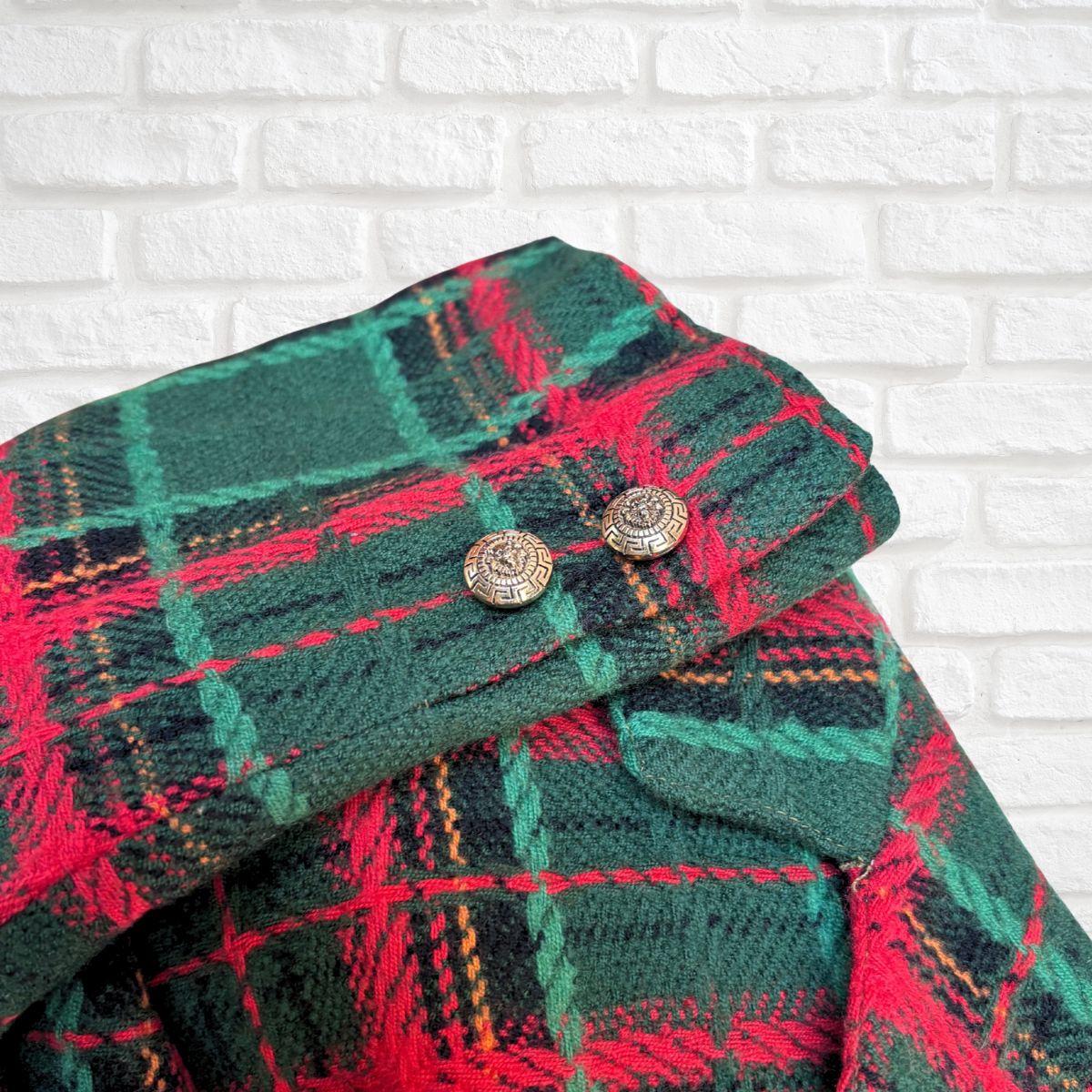 A close-up of a folded jacket in green, red, and black tartan with two round metallic buttons, set against a white brick wall background.