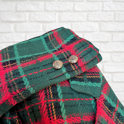 A close-up of a folded jacket in green, red, and black tartan with two round metallic buttons, set against a white brick wall background.
