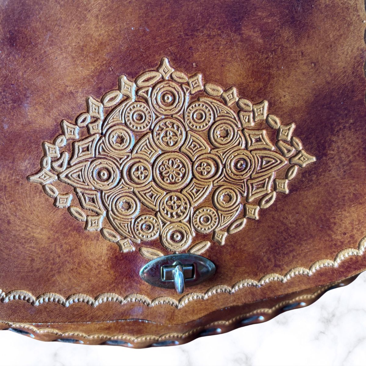 Close-up of a brown leather bag with an embossed ornate medallion pattern and a small metal clasp, photographed against a light marble-like background 