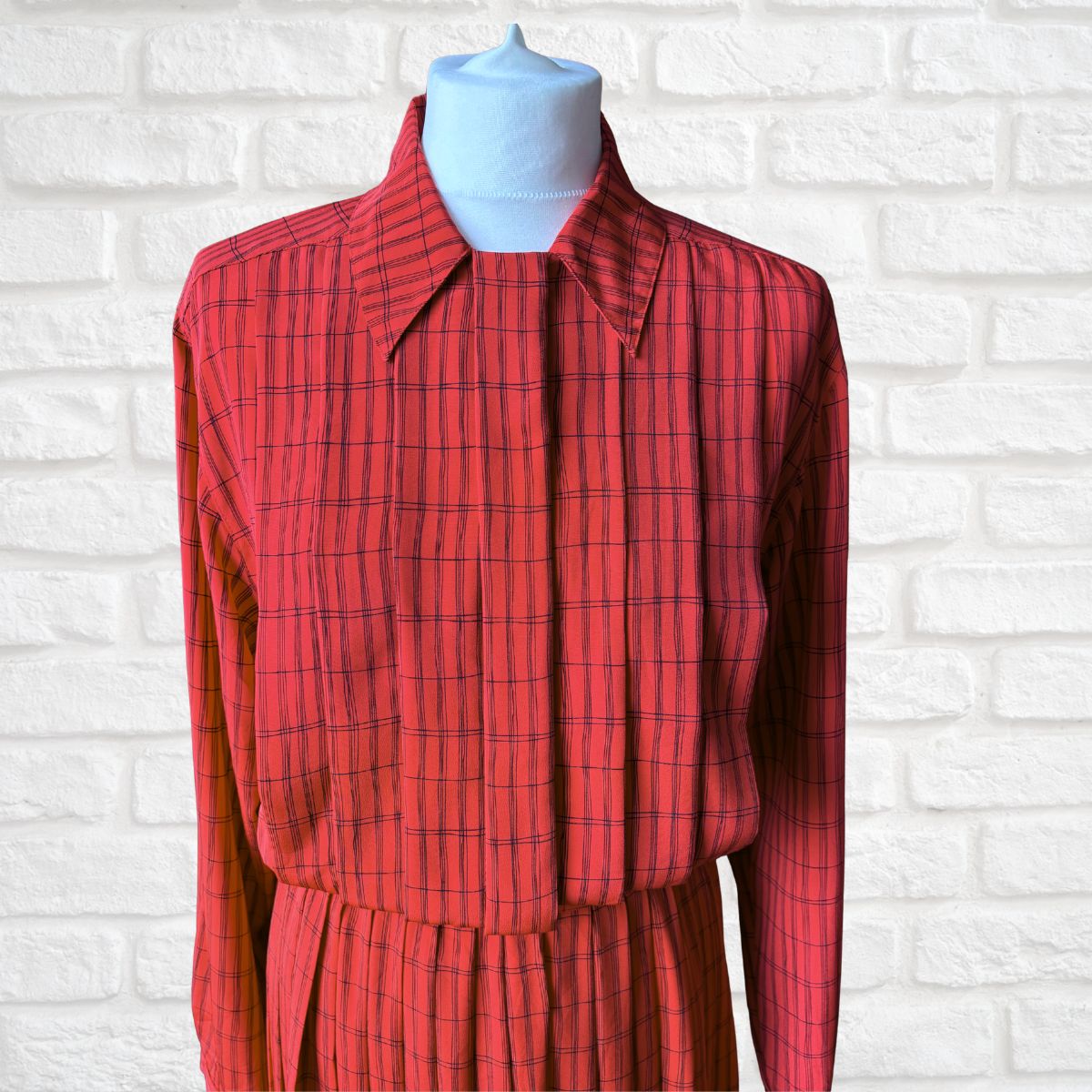 A red long-sleeve collared shirt dress with a thin black windowpane plaid pattern, displayed on a light blue mannequin torso against a white brick wall background.