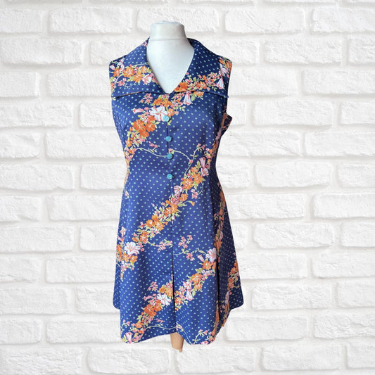 A sleeveless collared dress displayed on a white mannequin torso against a white brick wall. The dress has a blue base with white polka dots and diagonal floral stripes in orange, pink, and yellow.