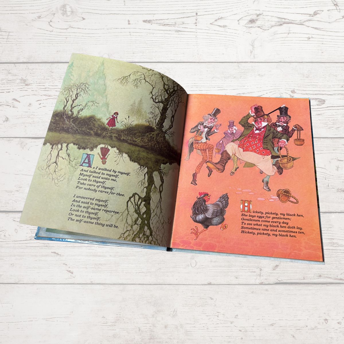 An open children's book spread on a light wood surface, showing illustrated pages with nursery rhyme text. The left page has a greenish forest scene with a red figure and the letter A, and the right page has an orange scene with characters and the letter H.